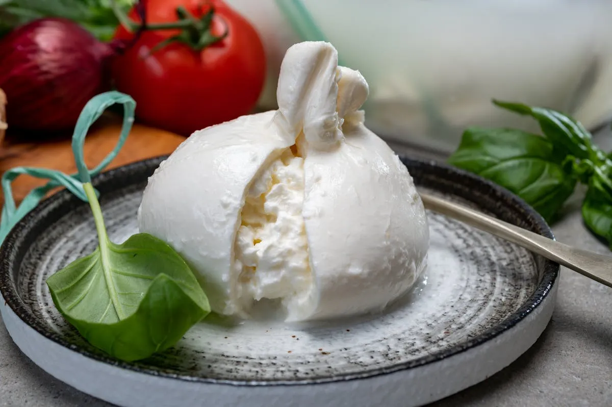 Plan rapproché d'une burrata découpée sur une assiette en céramique, cœur crémeux qui s'écoule, tomates cerises et basilic